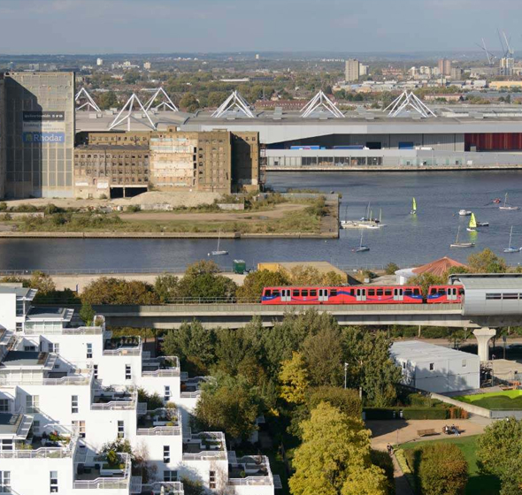 An aerial view of a DLR train crossing a bridge near riverside apartments, with cable car towers and industrial buildings in the background.