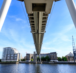 View from beneath a suspension footbridge over the water, with waterfront buildings and a crane in the distance.