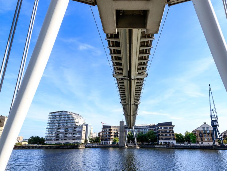 View from below a suspension footbridge over water, with waterfront buildings and a dockside crane in the background.