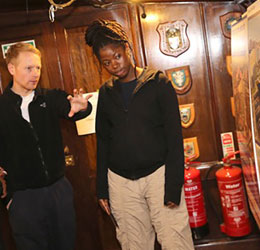 Man showing someone a display. Wooden walls with two fire extinguishers and some shield decoration.