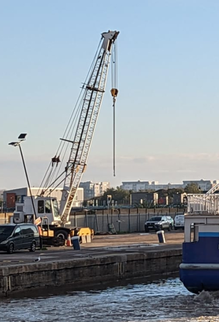 A mobile crane stands on a dockside, with vehicles and a boat nearby.