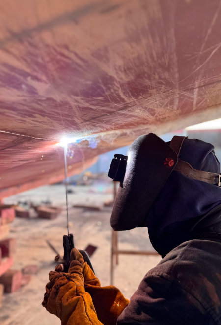 A welder in a protective mask and gloves works on dock edge repairs, with sparks flying from the weld.