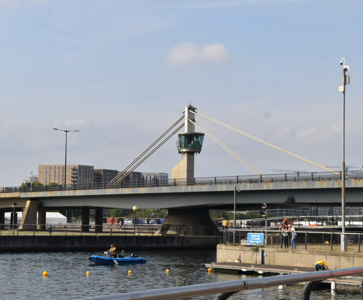 Connaught Bridge with its control tower spans Connaught Cut at Royal Albert Dock, with a small boat on the water below.