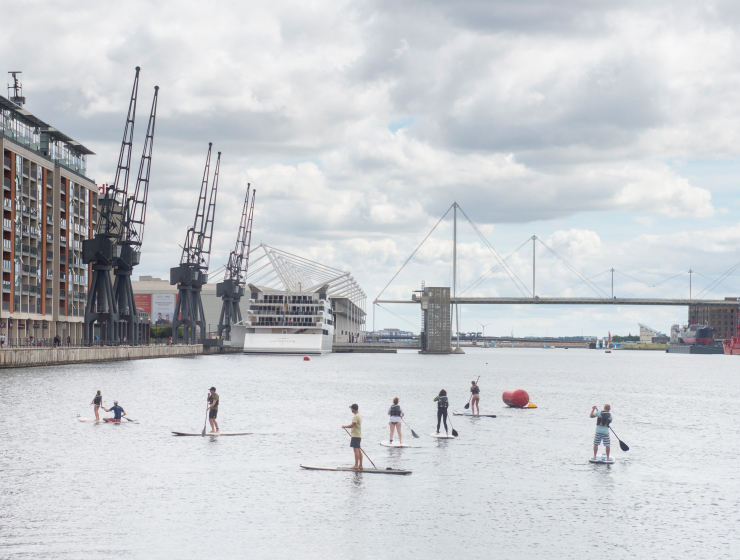 Paddleboarders on the water at Royal Docks, London, with dockside cranes and a footbridge in the background.