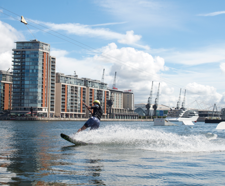 A cable wakeboarder on the dock, waterfront buildings and cranes behind.